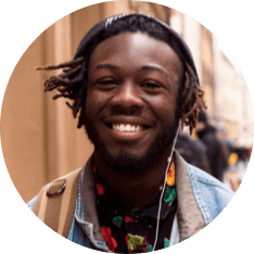 A person with a beard and dreadlocks smiles while wearing a beanie and denim jacket, walking outdoors.